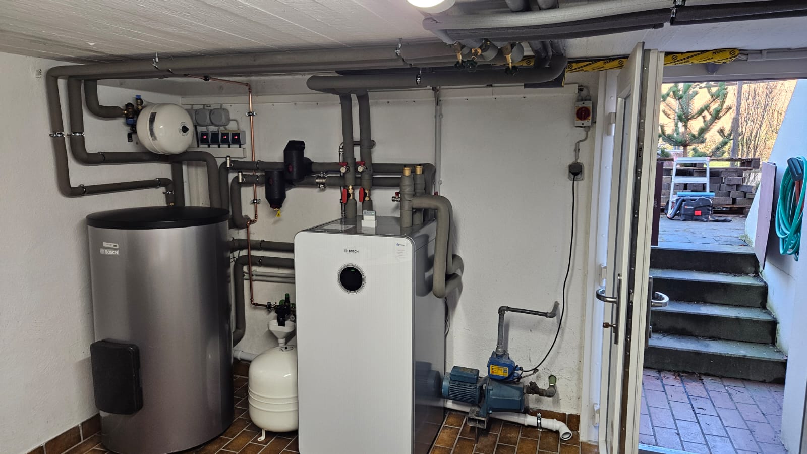 Im Heizungsraum steht eine Bosch-Wärmepumpe mit Pufferspeicher und gut isolierten Rohrleitungen, nahe einem Zugang zum Außenbereich.
